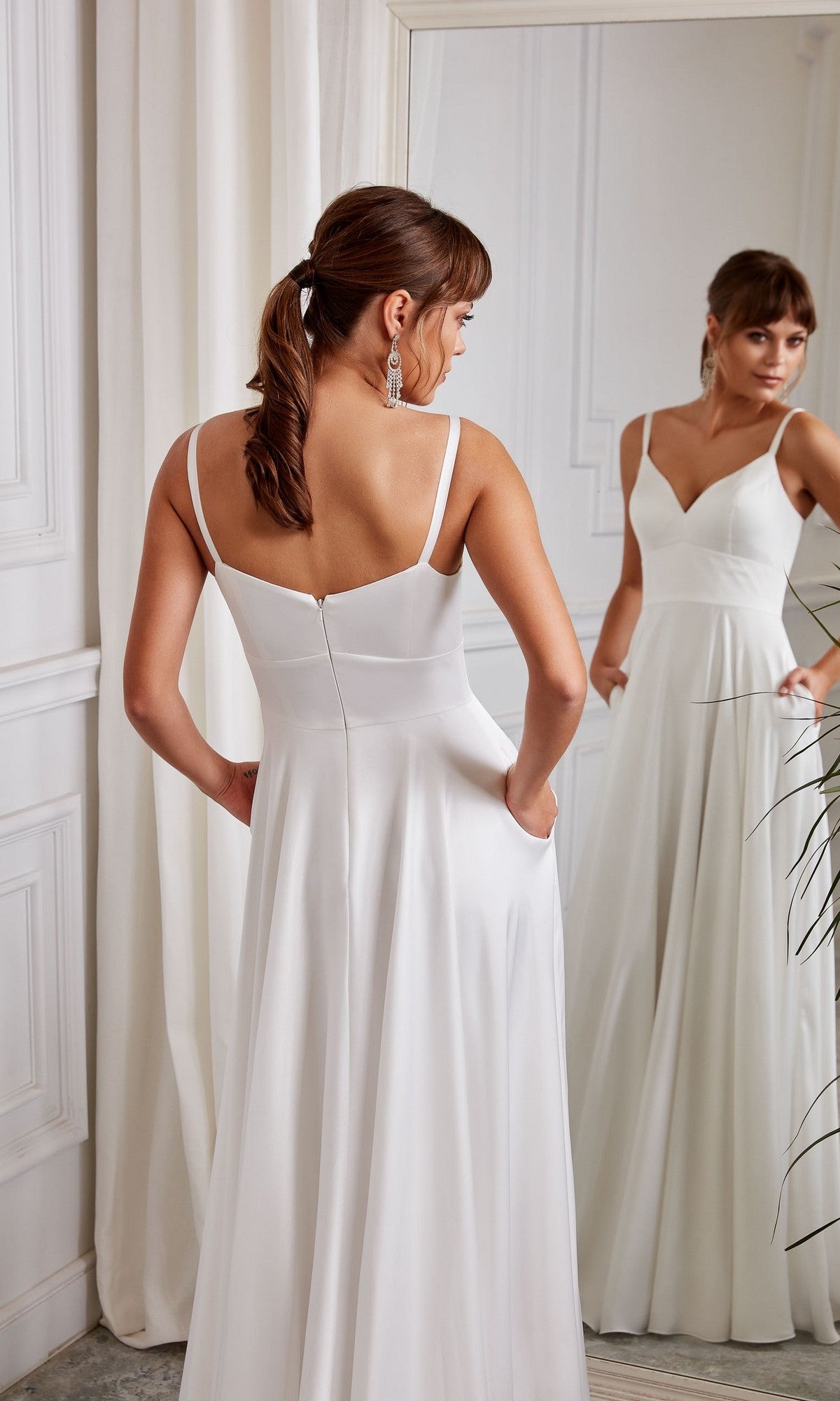 In a white-walled room, a woman in the Aerie Couture 2003 Long White Dress with an elegant v-neckline stands with her back to the camera, gazing at her reflection in a mirror.